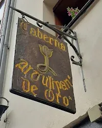 Letrero de madera colgado de un brazo de forja con las letras doradas Taberna Al Tulipán de Oro y un tulipán en el centro.
